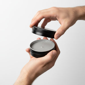 Person opening a black tin containing a texture paste on a light gray background